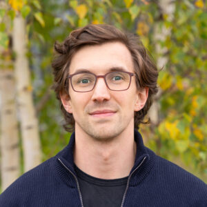 a portrait of Urs Spindler, with curly brown hair, a navy blue troyer sweater and square glasses.
