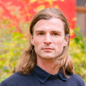 a portrait of Malte Spindler wearing long blonde hair and a navy blue pullover with a visible shirt collar.