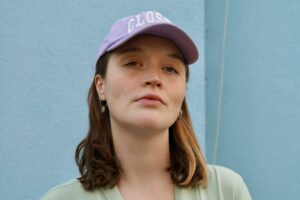 a portrait of Lara Gahlow, she wears a light blouse and pink cap with dark curly hair falling on her shoulders