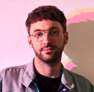 a portrait of Marcel Wicker he wears short dark hair and a full beard, an open shirt and glasses with rounded edges.
