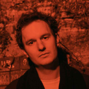a portrait of Timo Durst with a dark red tint, he wears a white shirt unter a dark jacked and short brown hair
