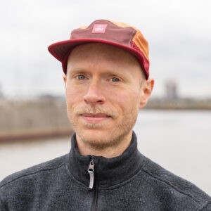 a portrait of Henning Walther wearing a dark sweater with a pulled-up zipper and a red cap on short-cropped blond hair