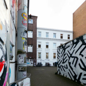 a photo looking through a graffiti bearing driveway on to a three story former factory building with a white classicist facade