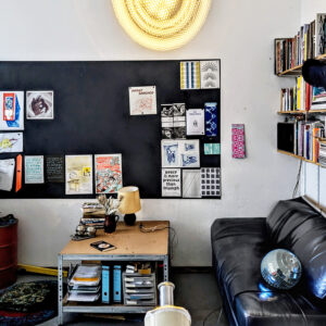 An image of the brueder coop office in Berlin, showing a blackboard with different colourful prints, a black leather couch and a book shelf and tables stacked with books.